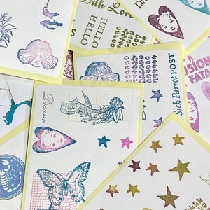 May include: A collection of art cards with various designs and text. The cards feature illustrations of faces, hearts, butterflies, and mermaids, along with text like "Dream," "Art Heals," and "With Love." The color palette includes shades of blue, pink, and gold.
