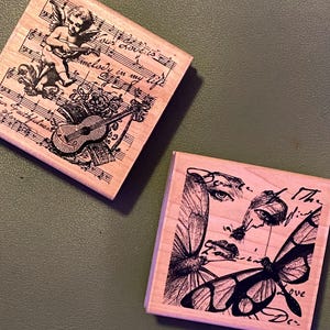 May include: Two square wooden stamps. One features an angel playing a guitar with musical notes and the text "Your love is melody in my life". The other stamp has a butterfly and a face with the text "The mind...love".