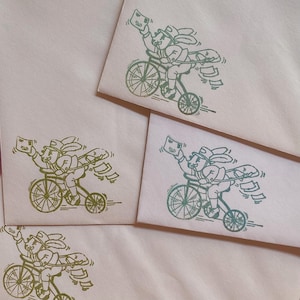 May include: Four light pink envelopes each featuring a green ink stamp illustration of a rabbit wearing a cap and carrying a mail bag while riding a bicycle. The rabbit is depicted holding an envelope aloft with one hand while letters fly.