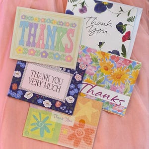 May include: A collection of floral-themed thank you cards. One card features the word "THANKS" in pastel colors. Other cards display "Thank You" and "Thank You Very Much" with floral and star designs. The cards are on a pink surface.