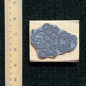 May include: A rubber stamp with a whimsical design of a person with sunflower-like features. The stamp is mounted on a rectangular wooden block. The ruler next to the stamp measures approximately 12.7 cm.