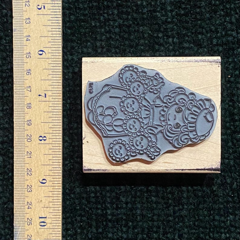 May include: A rubber stamp with a whimsical design of a person with sunflower-like features. The stamp is mounted on a rectangular wooden block. The ruler next to the stamp measures approximately 12.7 cm.