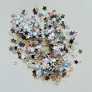 May include: A pile of star-shaped confetti cut from paper. The confetti is mainly white with printed text, and some stars have colourful accents. The stars are scattered on a white surface.