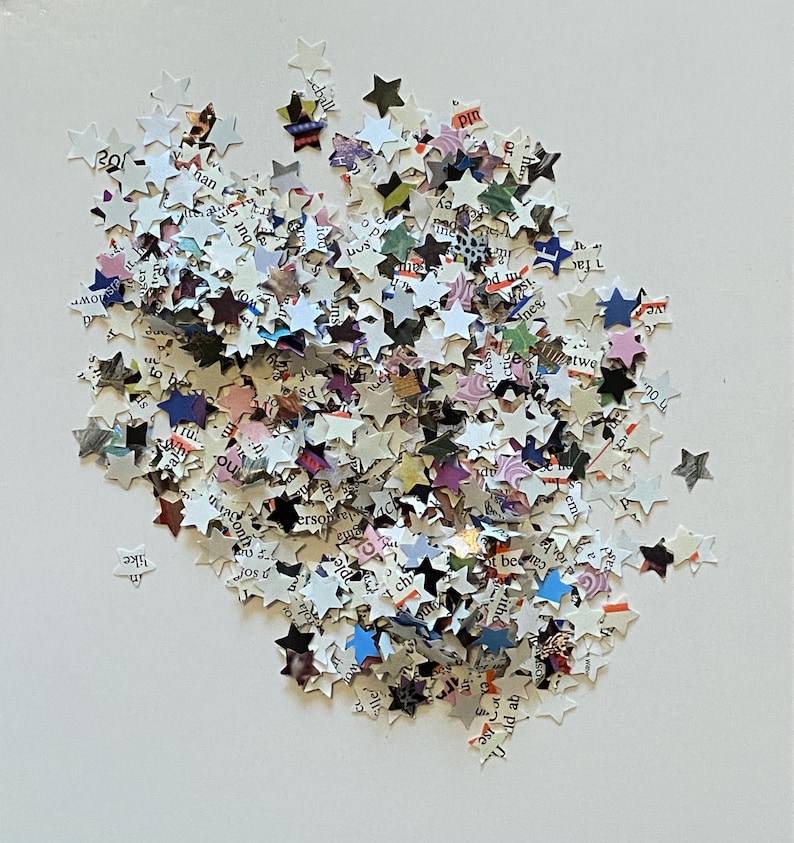 May include: A pile of star-shaped confetti cut from paper. The confetti is mainly white with printed text, and some stars have colourful accents. The stars are scattered on a white surface.