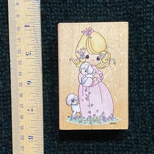 May include: A wooden stamp featuring a cartoon girl in a pink dress holding a white poodle. The girl has blonde hair and is adorned with flowers. A second poodle stands nearby, with floral accents at the base. The stamp measures approximately 7.6 cm in height.