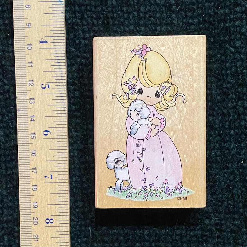 May include: A wooden stamp featuring a cartoon girl in a pink dress holding a white poodle. The girl has blonde hair and is adorned with flowers. A second poodle stands nearby, with floral accents at the base. The stamp measures approximately 7.6 cm in height.