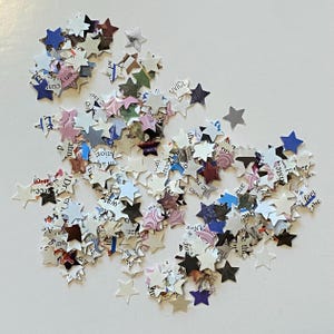 May include: A scattering of star-shaped confetti in shades of white, blue, and purple. The confetti is made from paper with printed text, giving it a distinctive look. Suitable for craft projects and decorations.
