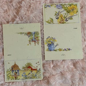 May include: Two cream-colored stationery sheets with floral and bird illustrations. One sheet features a birdhouse, a red barn, and blue flowers. The other has a watering can with flowers and a sunflower design. The word "STAMP" is printed on both.
