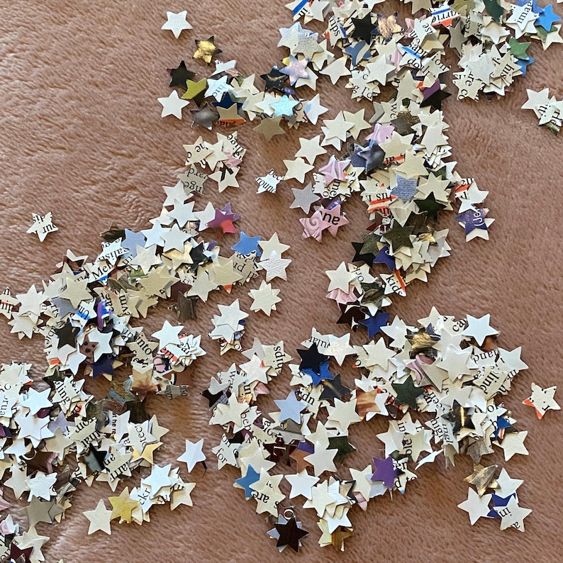 May include: A scattering of small, star-shaped confetti pieces on a pink surface. The stars are made from paper with printed text and various colours, including white, black, blue, and yellow. Suitable for crafts or decorations.