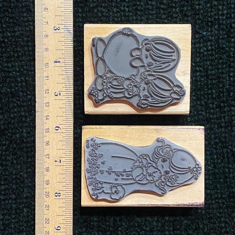 May include: Two rubber stamps with floral and character designs. The stamps are mounted on rectangular wooden blocks. One stamp features a character with a large head and floral accents. The other stamp shows a character lying down with floral details.