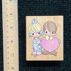 May include: A wooden stamp with a cartoon boy and girl holding a pink heart. The girl has blonde hair with blue bows, and the boy has brown hair and a yellow bow tie. The stamp measures approximately 5 cm by 5 cm.