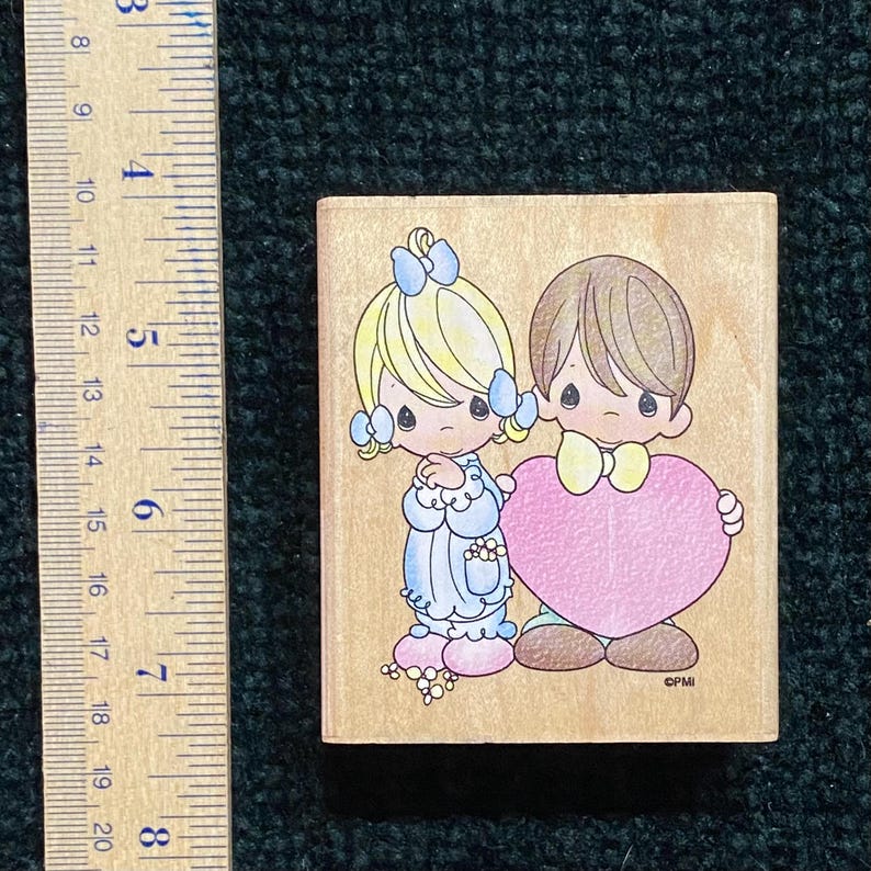 May include: A wooden stamp with a cartoon boy and girl holding a pink heart. The girl has blonde hair with blue bows, and the boy has brown hair and a yellow bow tie. The stamp measures approximately 5 cm by 5 cm.