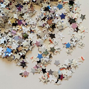 May include: A collection of star-shaped confetti pieces made from paper. The confetti is mainly white, with text and colourful accents. The stars vary in size and are scattered across a white surface, creating a festive appearance.