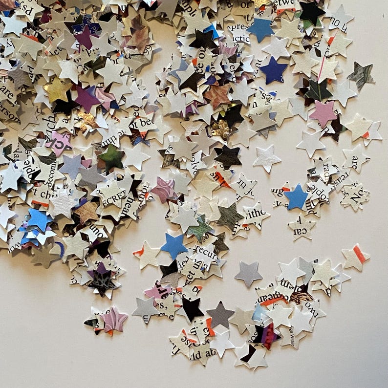 May include: A collection of star-shaped confetti pieces made from paper. The confetti is mainly white, with text and colourful accents. The stars vary in size and are scattered across a white surface, creating a festive appearance.