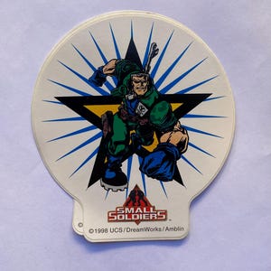 May include: A white sticker with a blue starburst background. A cartoon soldier in green and blue is standing in the center of a black star. The text "SMALL SOLDIERS" is at the bottom of the sticker with the copyright information "&copy; 1998 UCS/DreamWorks/Amblin".