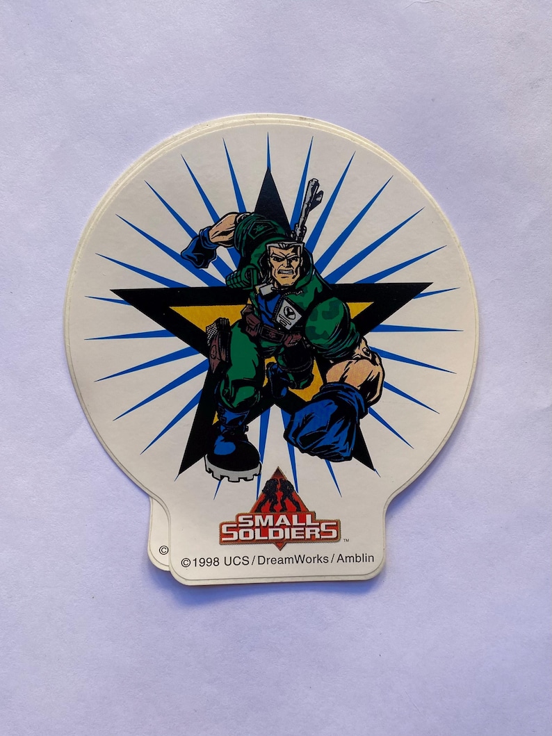 May include: A white sticker with a blue starburst background. A cartoon soldier in green and blue is standing in the center of a black star. The text "SMALL SOLDIERS" is at the bottom of the sticker with the copyright information "&copy; 1998 UCS/DreamWorks/Amblin".