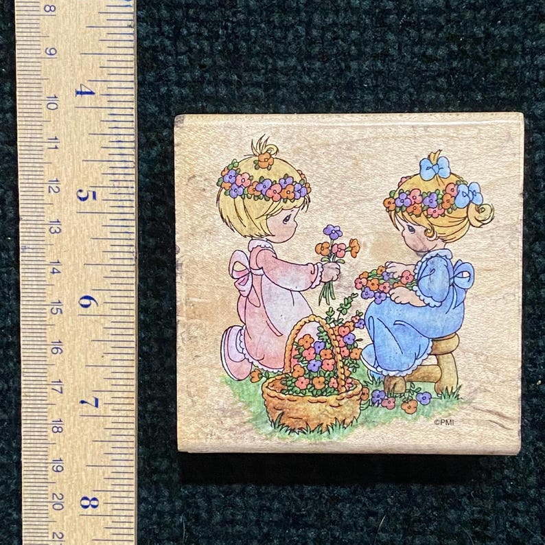 May include: A wooden stamp depicting two cartoon girls with blonde hair and floral crowns. One girl, in a pink dress, is holding flowers, while the other, in a blue dress, is seated. A basket of flowers is also present.