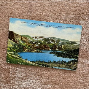 May include: Vintage postcard featuring a scenic view of Twin Lakes in the Beartooth Mountains, Montana. The postcard shows a blue lake surrounded by green trees and mountains with snow-capped peaks. Text on the top reads: "Twin Lakes in the Beartooth Mountains on Hi-Way 12 North East Entrance to Yellowstone National Park via Red Lodge, Montana."