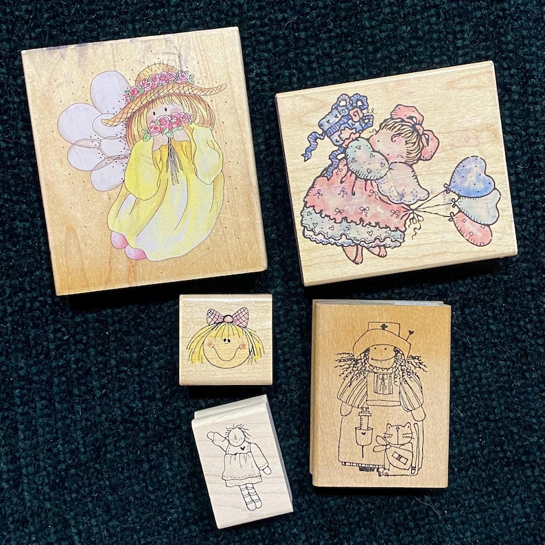 以下が含まれることがあります： 気まぐれなイラストが特徴の木製スタンプのコレクション。スタンプは、妖精、風船を持った女の子、リボンが付いた笑顔、人形、猫と看護師を描いています。イラストはソフトカラーと黒インクです。
