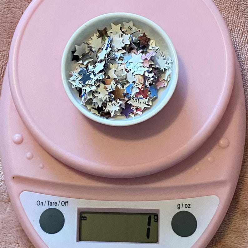 May include: A pink digital kitchen scale with a white bowl of star-shaped paper confetti. The scale shows a weight of 1 g. The bowl is centered on the scale, which has an On/Tare/Off button.
