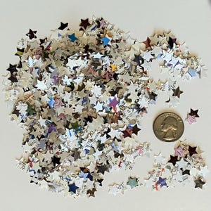 May include: A collection of small, star-shaped confetti pieces made from book pages. The confetti is mainly white with text and colourful accents. A coin is next to the confetti for size comparison.