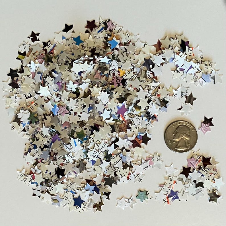 May include: A collection of small, star-shaped confetti pieces made from book pages. The confetti is mainly white with text and colourful accents. A coin is next to the confetti for size comparison.