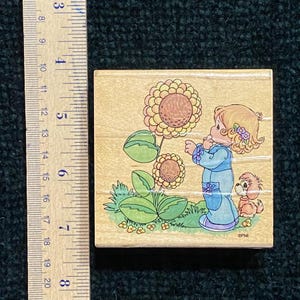 May include: A wooden stamp with a cartoon illustration of a girl in a blue dress looking at a sunflower. A small dog is next to her. The stamp is approximately 5 cm square.