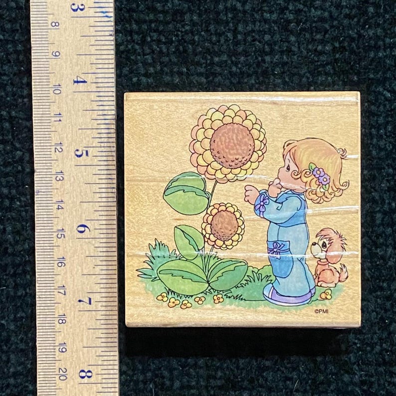 May include: A wooden stamp with a cartoon illustration of a girl in a blue dress looking at a sunflower. A small dog is next to her. The stamp is approximately 5 cm square.