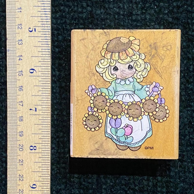 May include: A rectangular wooden stamp featuring a cartoon girl with blonde curly hair and a sunflower hat, holding smiling sunflowers. The girl wears a green top and white apron. The stamp has a light brown background.