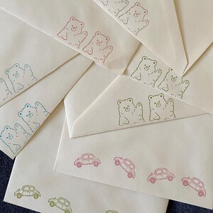 May include: A collection of cream-colored paper envelopes featuring stamped illustrations. Some envelopes display pairs of bears waving, while others feature rows of small cars. The stamps are rendered in various colors including red, blue, and green.