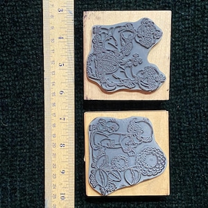 May include: Two rubber stamps with detailed floral and bird designs mounted on wooden blocks. The stamps are gray and the blocks are light brown. A ruler is placed next to the stamps, showing measurements in centimetres.