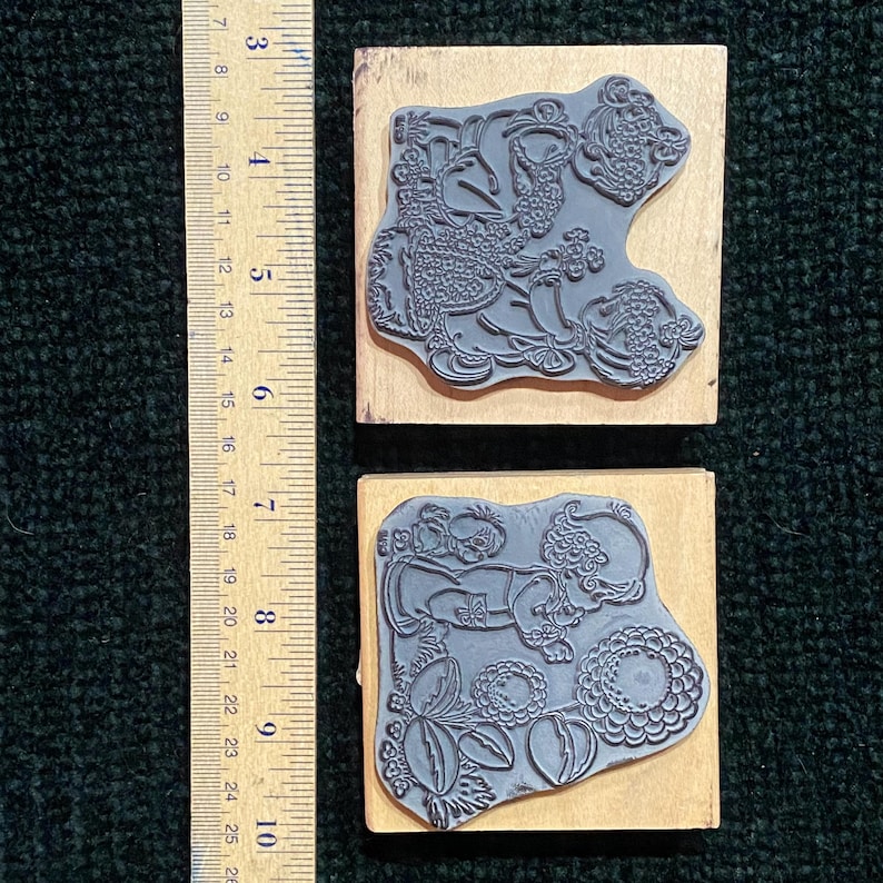 May include: Two rubber stamps with detailed floral and bird designs mounted on wooden blocks. The stamps are gray and the blocks are light brown. A ruler is placed next to the stamps, showing measurements in centimetres.