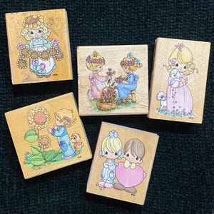 May include: Five wooden stamps with Precious Moments illustrations. The stamps show children with flowers, animals, and hearts in pastel colours. The stamps are on a dark green textured surface.