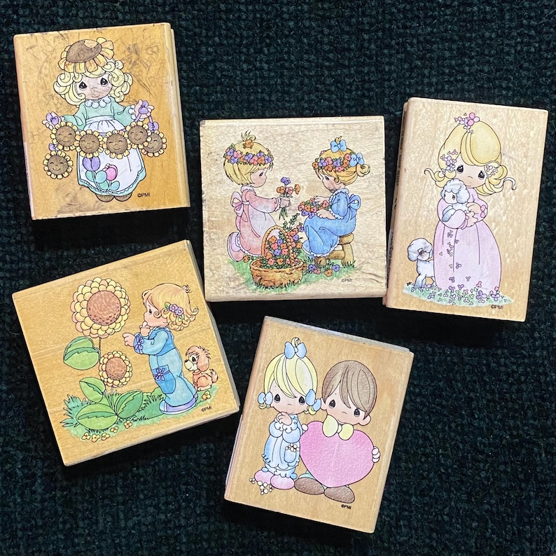 May include: Five wooden stamps with Precious Moments illustrations. The stamps show children with flowers, animals, and hearts in pastel colours. The stamps are on a dark green textured surface.