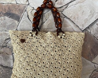 May include: A beige crocheted tote bag with a wooden handle. The bag has a woven pattern and a small brown tag with the word "Shop" on it.