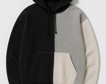 May include: A color-blocked hoodie in black, gray, and beige. The hooded sweatshirt features a kangaroo pocket and a drawstring. The design is split vertically, with each side in a different color. The hoodie is made of a soft, comfortable material.