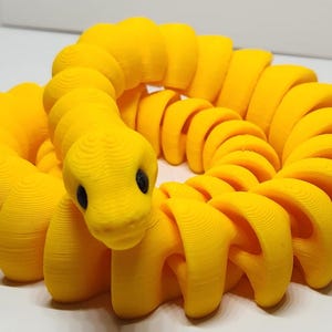 May include: A bright yellow, articulated snake toy with black eyes. The toy is made of connected, curved segments, allowing it to twist and turn. The snake is coiled, creating a playful and engaging appearance. The toy is likely 3D-printed.