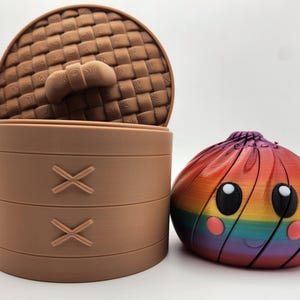 May include: A light brown cylindrical container with a woven-texture lid and two X-shaped embossed details on the side, positioned next to a round, rainbow-colored dumpling-shaped object featuring large black eyes and small pink circular cheeks.