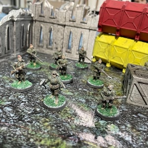 May include: A group of miniature figures dressed in military uniforms, holding rifles and standing on circular bases. The figures are arranged in a semi-circle on a textured surface.