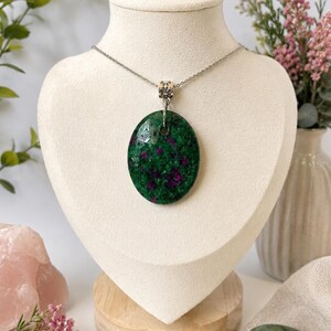 May include: An oval-shaped pendant featuring a polished green stone with deep magenta inclusions, suspended from a thin silver-toned metal chain. The pendant is attached to the chain by a small, patterned silver-toned metal bail at the top centre.