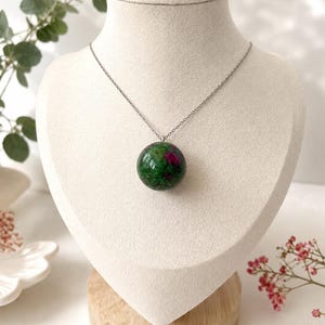 May include: A spherical pendant necklace featuring a polished green zoisite stone with vibrant magenta ruby inclusions. The smooth, round gemstone is suspended from a thin, silver-toned metal chain, creating a simple and elegant jewellery piece.