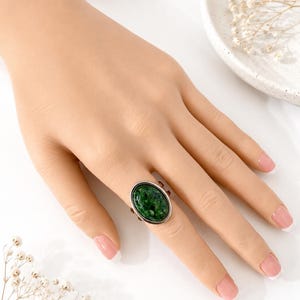 May include: A silver-toned ring featuring a large, oval-shaped cabochon with a deep green, mottled, and textured surface. The ring band is simple and metallic, showcasing a polished finish that complements the dark green stone setting.