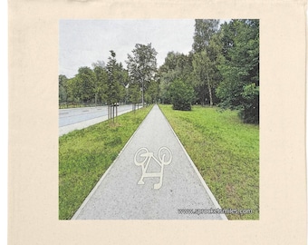 May include: A cream-coloured tote bag displaying a scenic image of a cycle path. The path is bordered by green grass, trees, and a road. A bicycle symbol is visible on the path. The bag has long, matching handles.