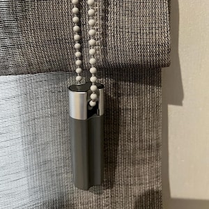 May include: Close-up of a window shade with a gray and silver color scheme. The shade is made of a textured fabric. A gray pull is attached to a beaded chain.