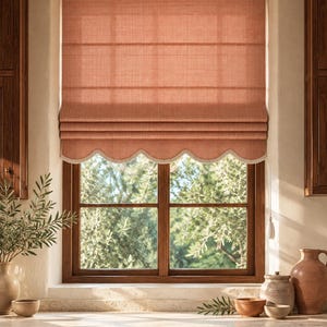 May include: A terracotta-colored Roman shade featuring a textured fabric finish and a scalloped bottom edge trimmed with white lace. The shade is mounted inside a window frame, partially covering the glass, and includes a white pull cord.