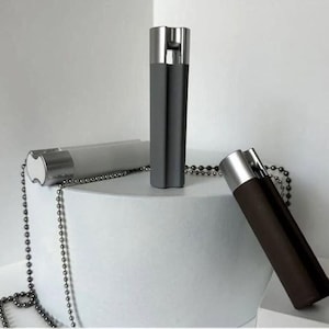 May include: Three cylindrical bottle stoppers with silver tops and dark bodies are displayed. One is gray, one is brown, and the third is a lighter gray. A silver beaded chain is draped around the stoppers.