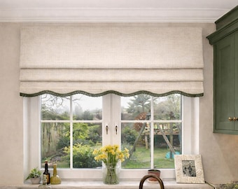 May include: A beige fabric roman shade with a scalloped bottom edge trimmed in dark green, installed over a window with white frames. The shade features a textured weave and hangs in horizontal folds above a kitchen sink area.