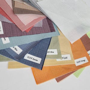 Op de afbeelding: Een verzameling stoffen stalen in verschillende kleuren, waaronder blauw, oranje en groen. Elk staal heeft een label met een kleurnaam, zoals "ELEN Denim" en "ELEN Orange". De stoffen hebben een geweven textuur.