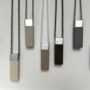 May include: Seven rectangular blind pulls in a range of colours: white, beige, taupe, brown, grey, and black. Each pull has a silver top and hangs from a beaded chain.