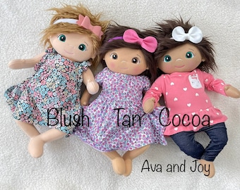 Puede incluir: Tres muñecas de tela hechas a mano con diferentes colores de pelo y atuendos. La muñeca de la izquierda lleva un vestido floral y tiene el pelo rubio. La muñeca del centro lleva un vestido floral y tiene el pelo castaño. La muñeca de la derecha lleva una camisa rosa con corazones blancos, pantalones vaqueros azules y tiene el pelo castaño. Las muñecas están etiquetadas como "Blush", "Tan" y "Cocoa".